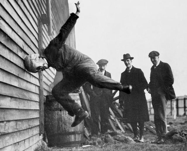 A group of men watching a man in the foreground leap head-first into a wall.